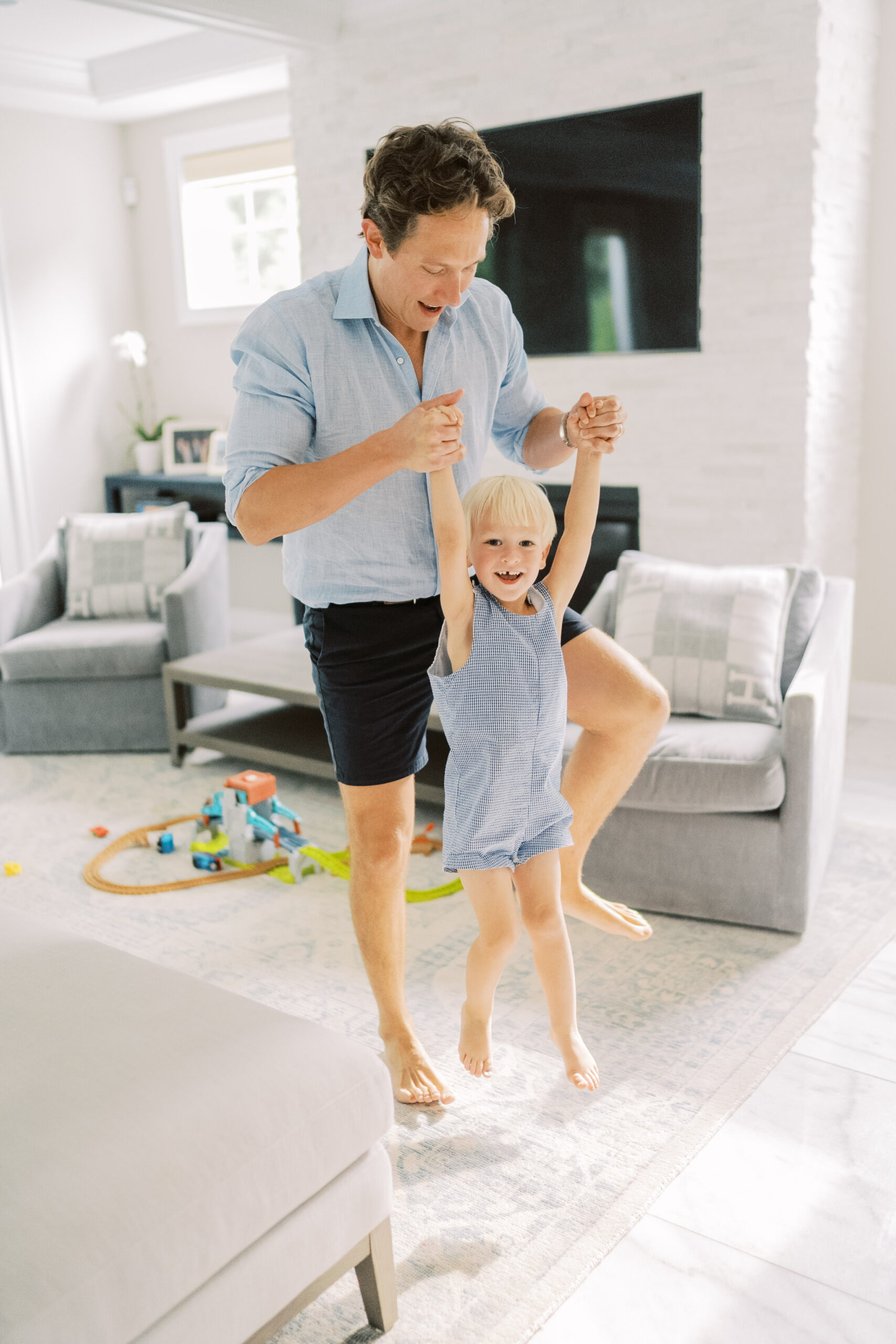 Dad swings his toddler son by his arms during their in home session. The son has a big smile on his face while he enjoys playing with his dad