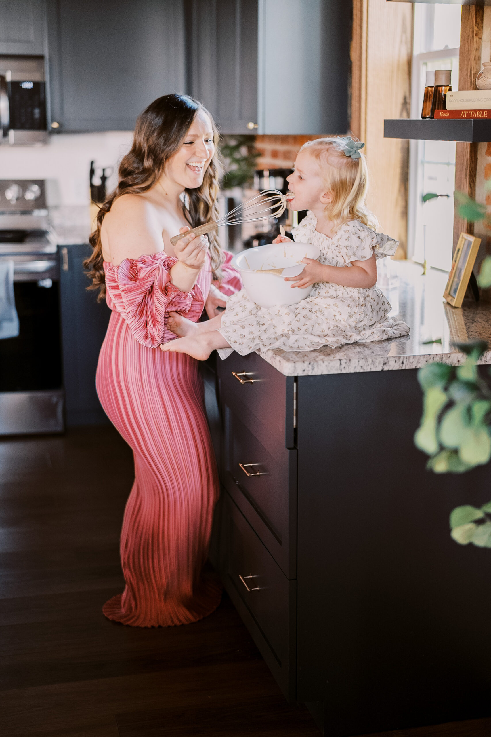 a mom in a long pink dress leans against the kitchen counter where her toddler daughter is sitting with a bowl of pancake batter. they are tasting the batter together during the Philadelphia Family Photographer Session.