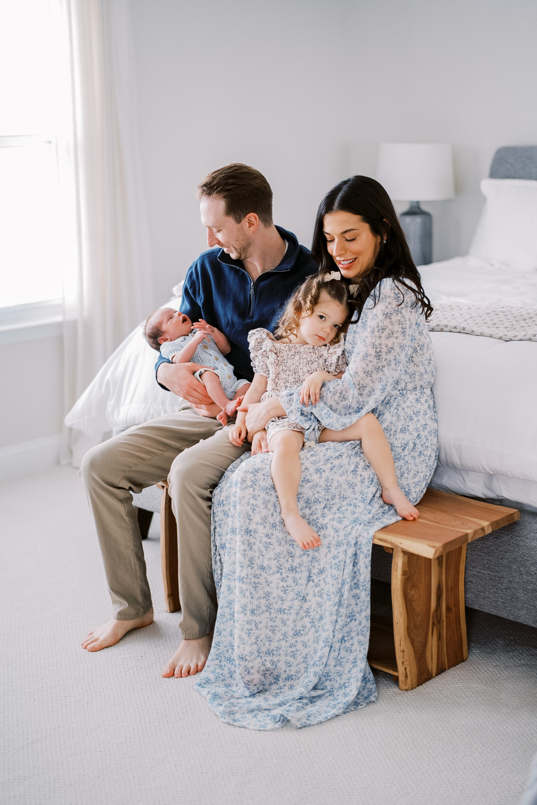 family is sitting on a bench in front of their bed. dad is craddling his newborn son, mom is holding her toddler daughter.