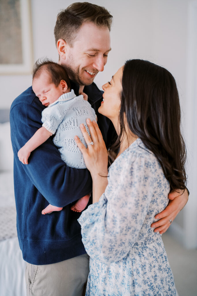 A mom and dad stand close while the dad has his newborn son on his shoulder