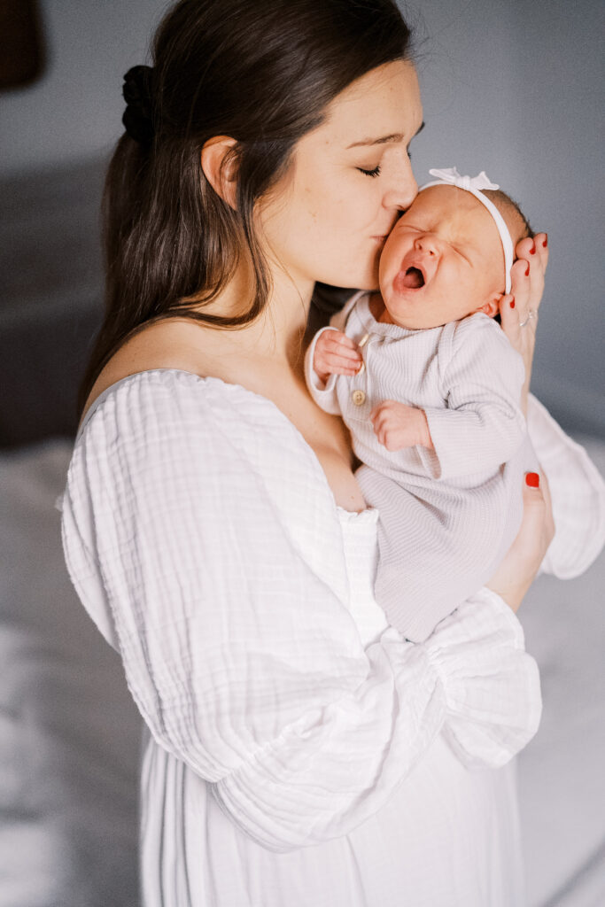 Woman stands in her bedroom kissing her yawning newborn on the cheek. She cuddles her baby during her Hershey Newborn Photography Session
