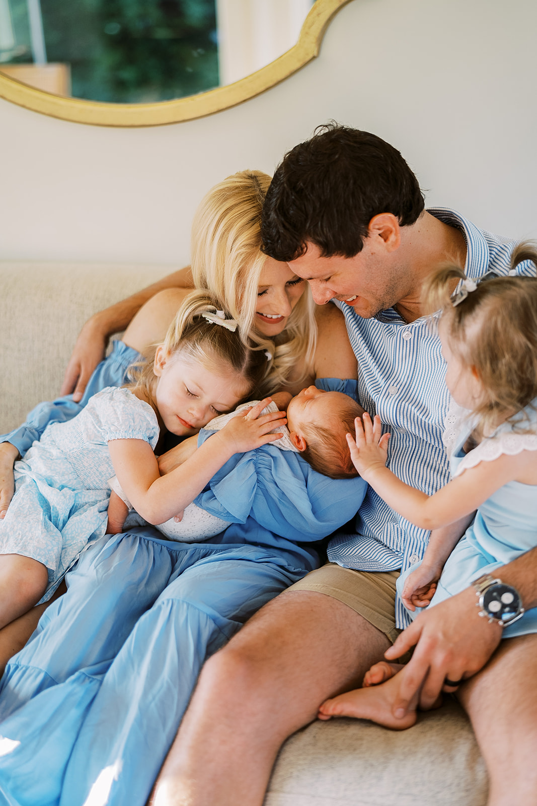 A family cuddles on their couch. The mom is holding her newborn baby boy, while her husband and two daughters crowd around in a big group hug during their Lancaster Newborn Photography session.