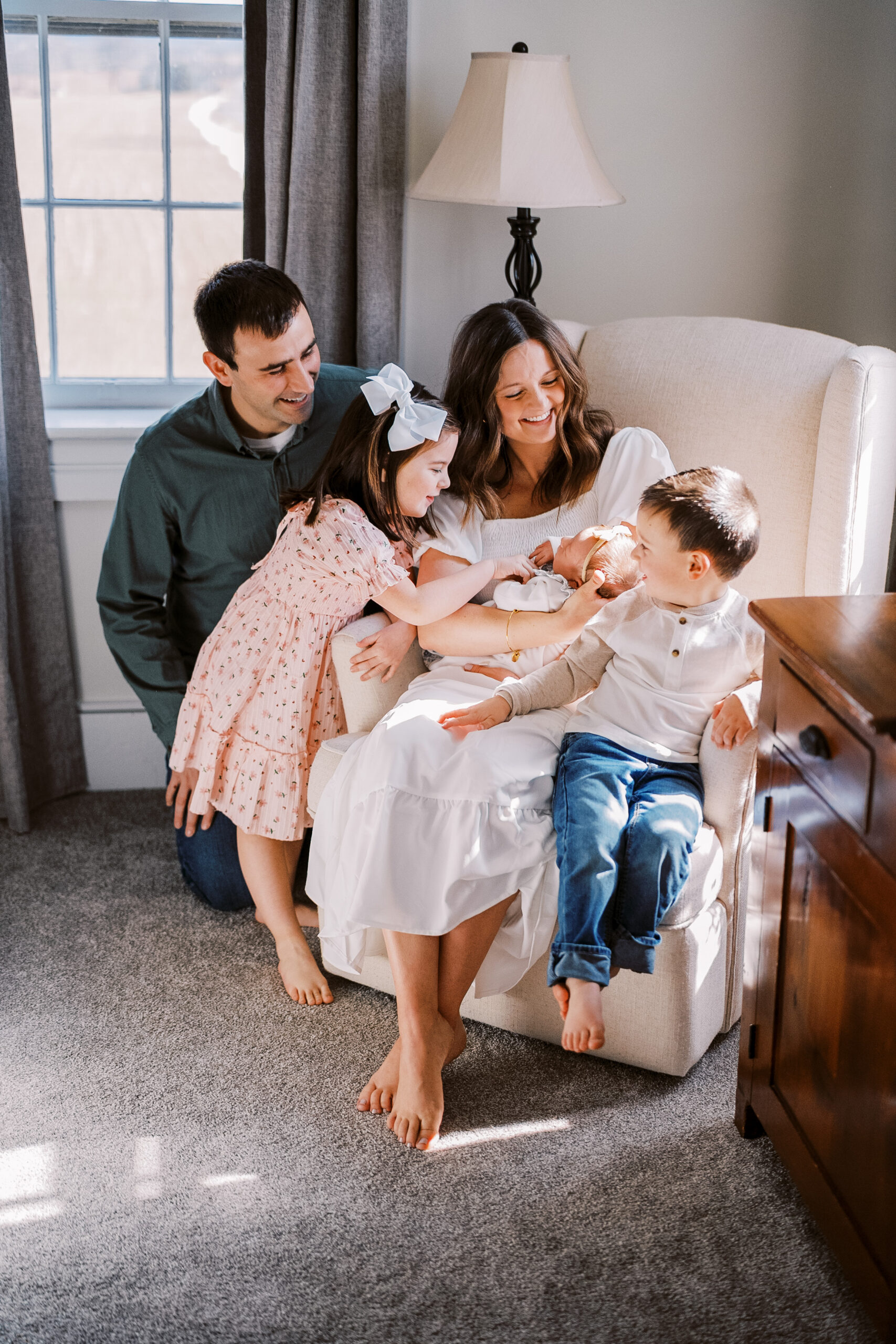 A family of 5 sits around the rocking chair while the mom holds a newborn baby on her lap. The family is smiling and happy during their newborn photography with family session