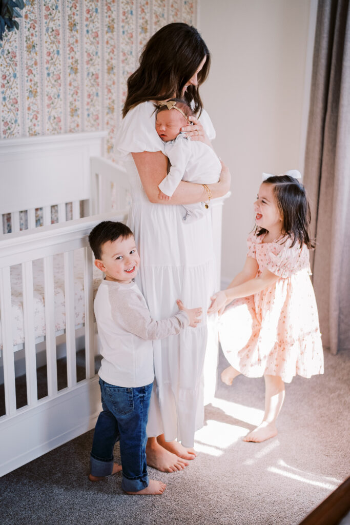 A mom stands in her baby girl's nursery holding her sleeping newborn baby against her chest. Her son and daughter on holding on to the skirt of her dress and laughing. The family is enjoying their newborn photography session.